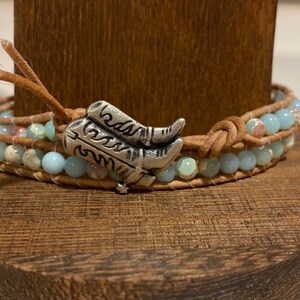 May include: A light blue and brown leather wrap bracelet with a silver boot charm. The bracelet is made with multiple strands of leather and beads.