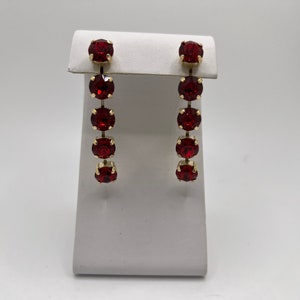 May include: A pair of red crystal dangle earrings with a gold setting. The earrings have five crystals each, arranged in a vertical line.