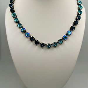 May include: A necklace with a gradient of blue and green crystals on a silver chain. The necklace is displayed on a white mannequin bust.