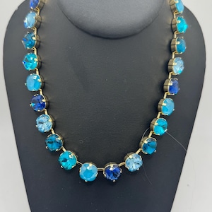 May include: A gold chain necklace with a gradient of blue round crystals. The necklace is on a black display stand.