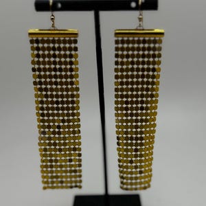 May include: A pair of gold chain earrings with a rectangular shape. The earrings are hanging from a black stand.