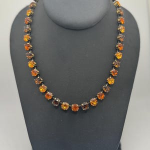 May include: A necklace featuring a single strand of round, faceted amber and orange-toned crystals set in a dark, metallic prong setting. The stones alternate in shades of deep brown, burnt orange, and bright yellow, creating a gradient effect.