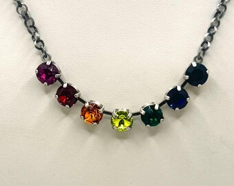 Puede incluir: Un collar de cadena plateada con un arcoíris de piedras preciosas de colores. Las piedras están dispuestas en una línea curva, con colores que van del morado al azul. El collar tiene un diseño sencillo y elegante.