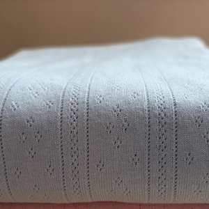 May include: Two folded pieces of fabric stacked on top of each other. The top piece is light blue and the bottom piece is light pink. Both fabrics feature a vertical ribbed texture with a repeating delicate eyelet pattern between the ribs.