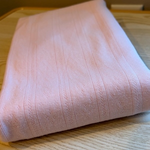 May include: A folded piece of light pink fabric featuring a subtle vertical ribbed texture and a delicate, repeating pointelle pattern between the ribs. The textile has a soft, knit appearance with a consistent pastel color throughout the entire surface.