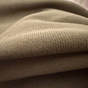 May include: A close-up view of a light tan, finely knit fabric with a consistent, repeating stitch pattern. The material displays soft folds and shadows, highlighting the texture of the knit construction across the surface of the textile.