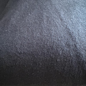 May include: A solid black textile fabric featuring a consistent vertical ribbed knit pattern. The material displays a dense, textured surface with fine fibers visible across the dark, uniform color, creating a structured and tactile appearance.