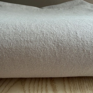 May include: A folded piece of light gray fabric with a visible ribbed knit texture. The material features a consistent vertical pattern of raised lines across the surface, creating a soft and uniform appearance throughout the textile.