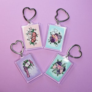 May include: Four keychains with different designs featuring video game controllers and flowers. Each keychain has a heart-shaped metal ring and a clear plastic charm with a colorful illustration.
