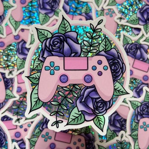 May include: A pink video game controller with purple flowers and green leaves on a holographic background. The controller has a heart-shaped button on each side.