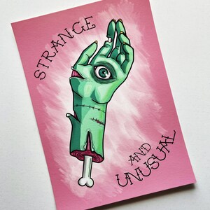 May include: A pink background with a green hand with an eye in the palm. The hand is holding a bone. The text "STRANGE AND UNUSUAL" is written in black on the pink background.