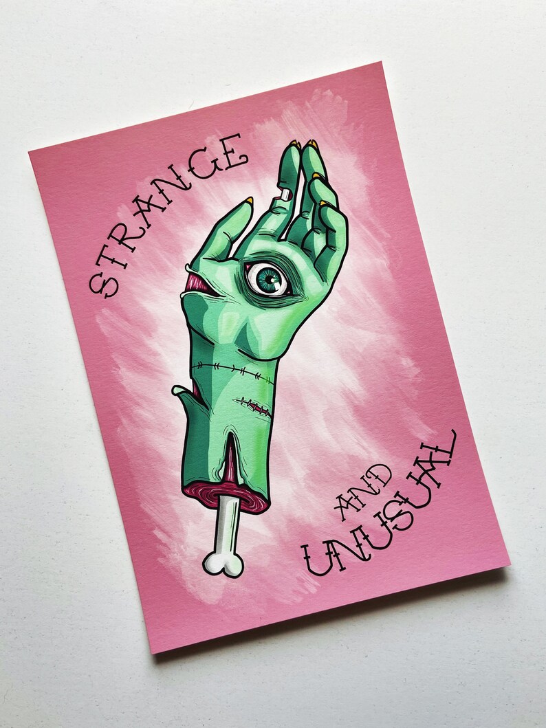 May include: A pink background with a green hand with an eye in the palm. The hand is holding a bone. The text "STRANGE AND UNUSUAL" is written in black on the pink background.