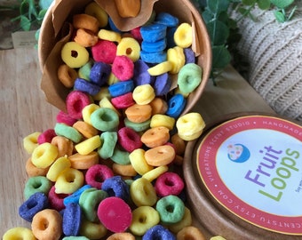 May include: A collection of colorful, ring-shaped wax melts resembling cereal, spilling from a small, overturned cardboard container. The melts are in various colors, including red, green, yellow, and blue. A wooden lid with the text "Fruit Loops" is also visible.