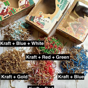 May include: Kraft paper gift boxes with clear windows, decorated with holiday-themed illustrations. The boxes are paired with shredded paper in various colors, including blue, white, gold, red, and green, with text labels.