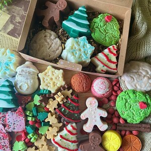 May include: A festive gift box filled with assorted Christmas-themed soaps and candies. The box contains gingerbread men, Christmas trees, snowflake shapes, and round soaps in various colors, including green, white, brown, and red. The arrangement is set on a neutral background.