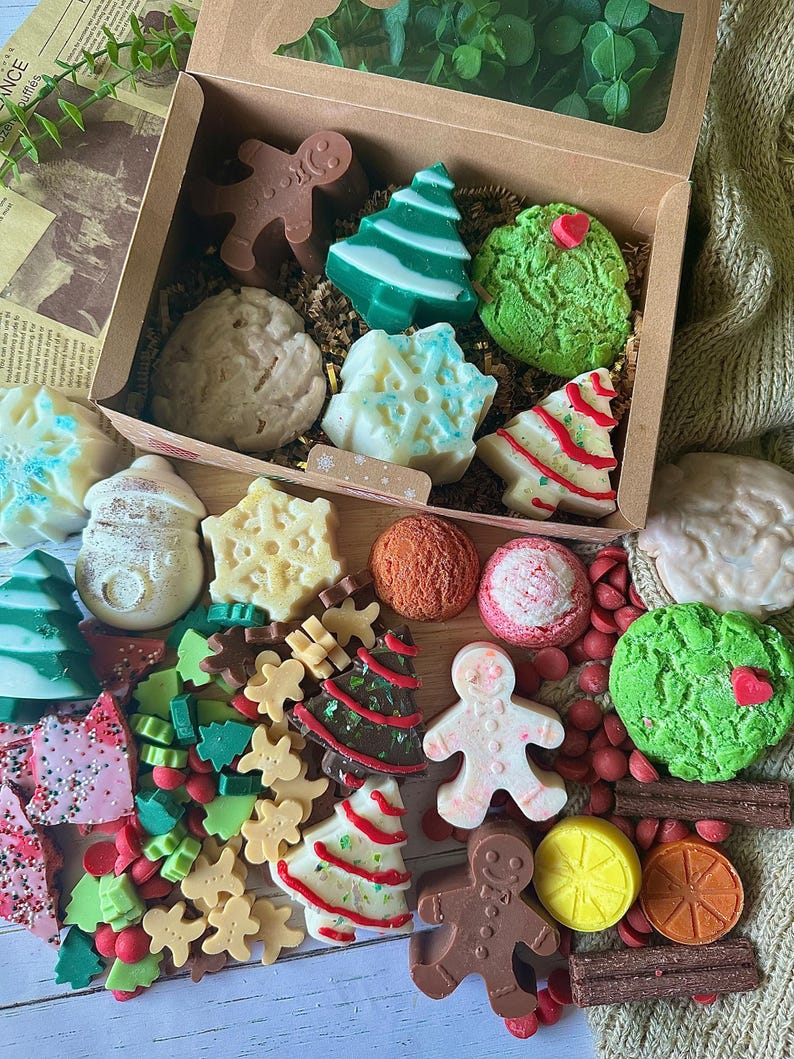 May include: A festive gift box filled with assorted Christmas-themed soaps and candies. The box contains gingerbread men, Christmas trees, snowflake shapes, and round soaps in various colors, including green, white, brown, and red. The arrangement is set on a neutral background.