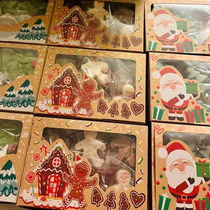 May include: Several Christmas-themed boxes filled with cookies. The boxes feature illustrations of Santa Claus, gingerbread houses, and snowmen. The cookies inside are various shapes, including gingerbread men, trees, and stars. The boxes are brown with clear windows.