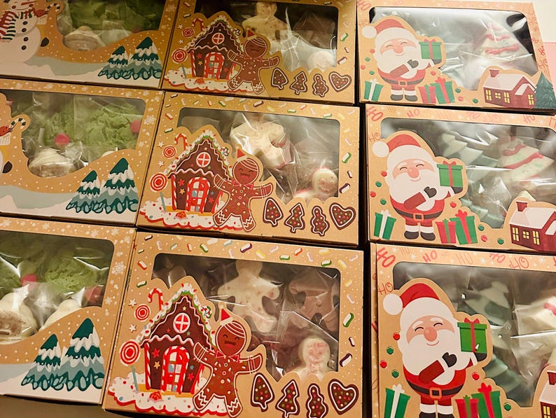 May include: Several Christmas-themed boxes filled with cookies. The boxes feature illustrations of Santa Claus, gingerbread houses, and snowmen. The cookies inside are various shapes, including gingerbread men, trees, and stars. The boxes are brown with clear windows.