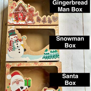 May include: Three festive cardboard boxes with clear windows. The top box features a gingerbread house and man, the middle box a snowman, and the bottom box Santa Claus. Each box has a holiday theme, with the text "Gingerbread Man Box", "Snowman Box", and "Santa Box".