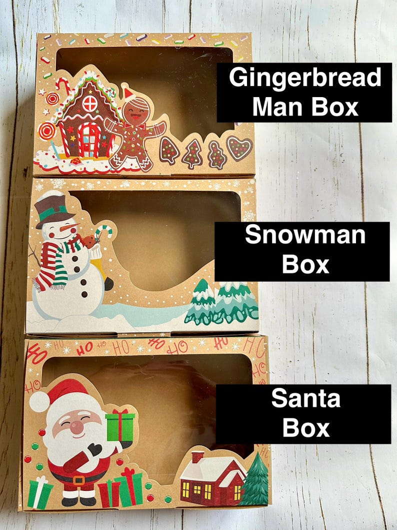 May include: Three festive cardboard boxes with clear windows. The top box features a gingerbread house and man, the middle box a snowman, and the bottom box Santa Claus. Each box has a holiday theme, with the text "Gingerbread Man Box", "Snowman Box", and "Santa Box".