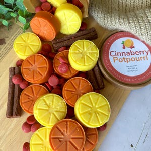 May include: A wooden cutting board displays a collection of wax melts and potpourri. The melts are shaped like orange and lemon slices, in yellow and orange hues. Red, round wax melts and brown stick-shaped melts are scattered around. A container reads "Cinnaberry Potpourri".