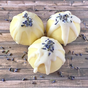 May include: Three round, light yellow soaps with white icing drizzled over the top and topped with dried lavender buds. The soaps are arranged on a wooden surface, with scattered lavender pieces.