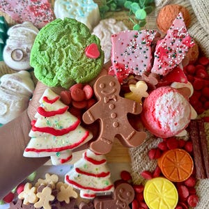 May include: A collection of festive, colorful soaps in various shapes, including a green tree, a gingerbread man, and a Christmas tree. The soaps are decorated with red and white accents and sprinkles, creating a holiday theme.