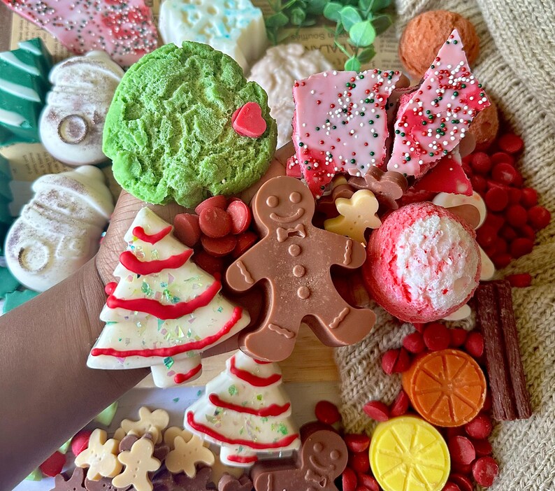 May include: A collection of festive, colorful soaps in various shapes, including a green tree, a gingerbread man, and a Christmas tree. The soaps are decorated with red and white accents and sprinkles, creating a holiday theme.