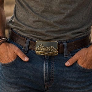 May include: A rectangular belt buckle featuring a raised, metallic mountain range relief design in a bronze tone, attached to a dark brown leather belt worn with blue denim jeans and a grey t-shirt, showcasing a detailed landscape scene.