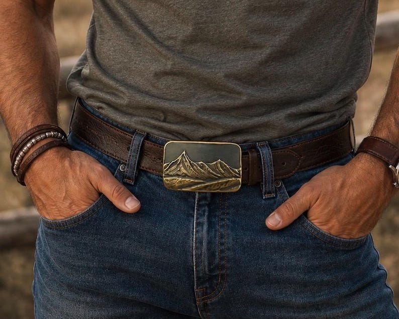 May include: A rectangular belt buckle featuring a raised, metallic mountain range relief design in a bronze tone, attached to a dark brown leather belt worn with blue denim jeans and a grey t-shirt, showcasing a detailed landscape scene.