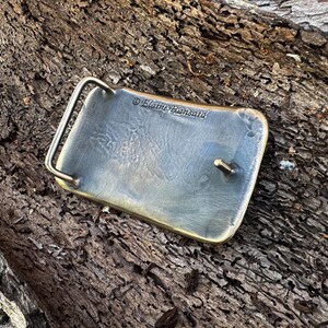 May include: A rectangular metal belt buckle with a brushed, weathered finish and rounded corners. The back features a hinged metal loop and a central prong for fastening. The text Elaine Hansard is engraved into the metal surface.