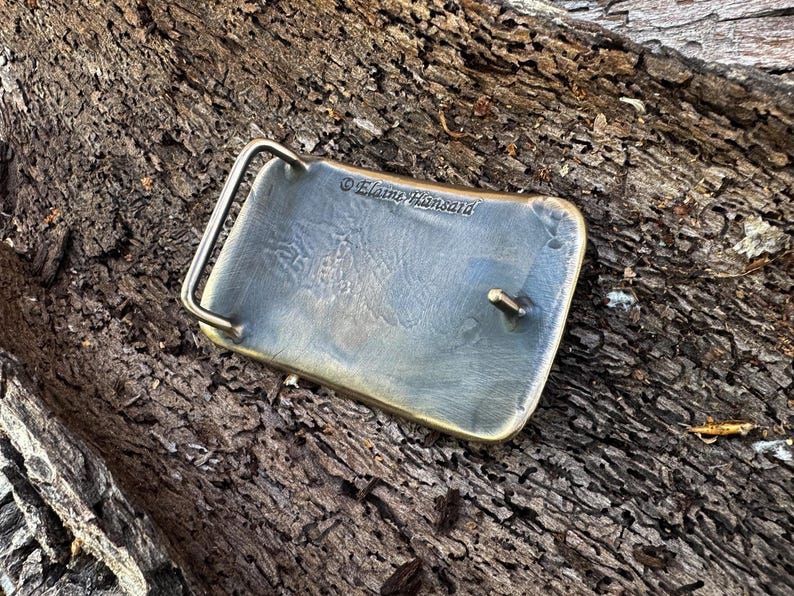 May include: A rectangular metal belt buckle with a brushed, weathered finish and rounded corners. The back features a hinged metal loop and a central prong for fastening. The text Elaine Hansard is engraved into the metal surface.