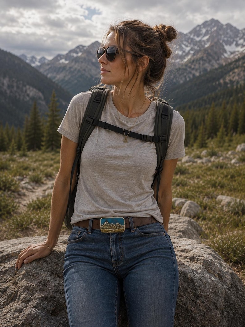 May include: A rectangular belt buckle featuring a raised gold-tone mountain range design against a teal blue enamel background, attached to a brown leather belt worn with blue denim jeans, a light gray t-shirt, and a dark gray backpack.