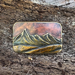 May include: A rectangular belt buckle made of brass and copper featuring a raised relief design of a mountain range. The sky area has a dark reddish-brown patina, while the mountains and foreground terrain display a polished golden brass finish.