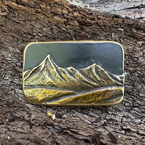 May include: A rectangular brass belt buckle featuring a raised, three-dimensional relief of a mountain range. The metal has a polished gold-tone finish on the peaks and slopes, contrasting with a dark, recessed background that highlights the texture.