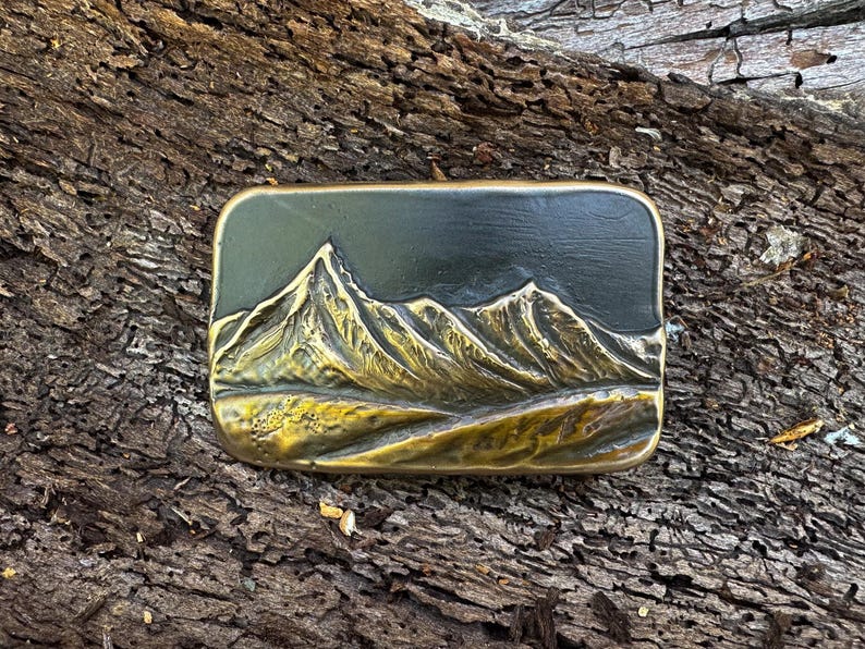 May include: A rectangular brass belt buckle featuring a raised, three-dimensional relief of a mountain range. The metal has a polished gold-tone finish on the peaks and slopes, contrasting with a dark, recessed background that highlights the texture.