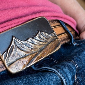 May include: A rectangular metal belt buckle featuring a raised, three-dimensional mountain range relief with a dark, oxidized finish. The buckle is attached to a tan leather belt with visible stitching, worn over blue denim jeans and a pink shirt.