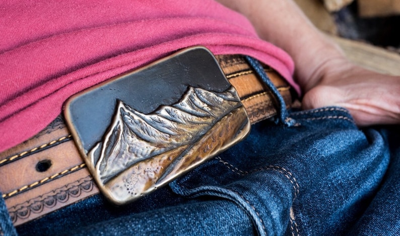 May include: A rectangular metal belt buckle featuring a raised, three-dimensional mountain range relief with a dark, oxidized finish. The buckle is attached to a tan leather belt with visible stitching, worn over blue denim jeans and a pink shirt.
