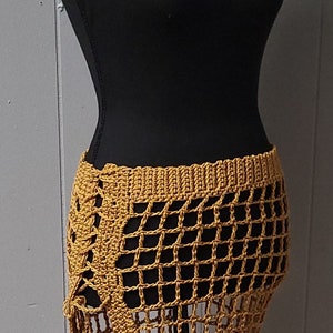 Puede incluir: Falda de crochet amarillo mostaza exhibida en un maniqu&iacute; negro. La falda presenta un dise&ntilde;o similar a una red, cintura alta y abertura lateral. El trabajo de crochet es detallado, mostrando la textura y el tejido abierto de la prenda.