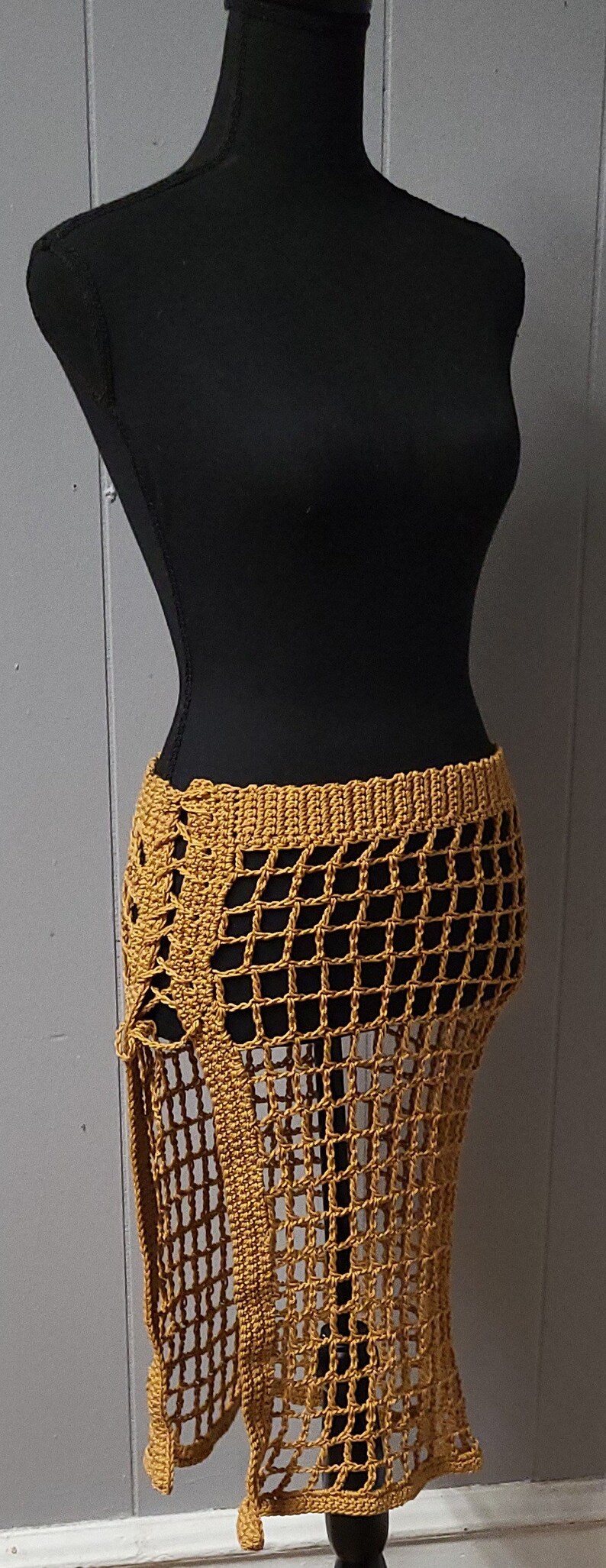 Puede incluir: Falda de crochet amarillo mostaza exhibida en un maniqu&iacute; negro. La falda presenta un dise&ntilde;o similar a una red, cintura alta y abertura lateral. El trabajo de crochet es detallado, mostrando la textura y el tejido abierto de la prenda.
