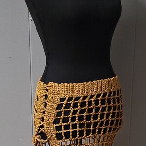 Puede incluir: Falda de ganchillo amarillo mostaza con un dise&ntilde;o de red. La falda se muestra en un maniqu&iacute; negro. La falda tiene una cintura alta y una abertura lateral. La falda est&aacute; hecha de un material suave y ligero.