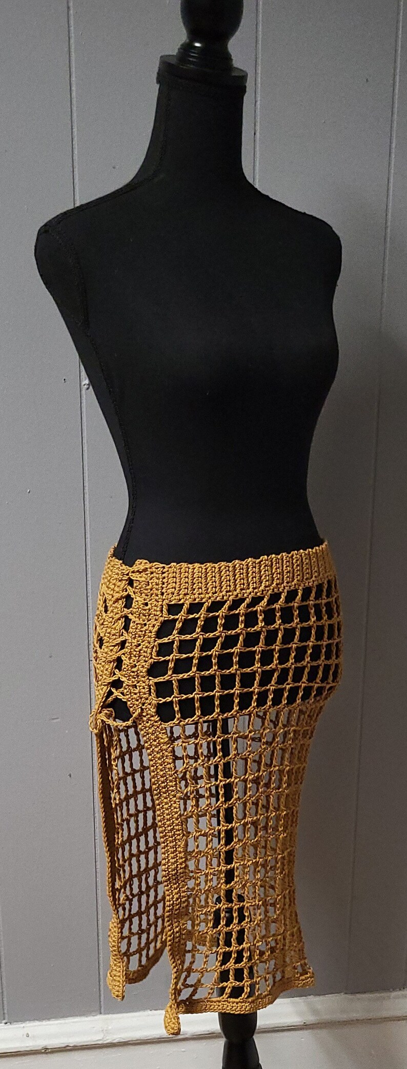 Puede incluir: Falda de ganchillo amarillo mostaza con un dise&ntilde;o de red. La falda se muestra en un maniqu&iacute; negro. La falda tiene una cintura alta y una abertura lateral. La falda est&aacute; hecha de un material suave y ligero.