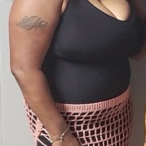 Puede incluir: Una mujer con una camiseta sin mangas negra y una falda de crochet rosa. La falda tiene un dise&ntilde;o de red. La mujer tiene el pelo rizado oscuro y un tatuaje en el brazo. Est&aacute; sonriendo.