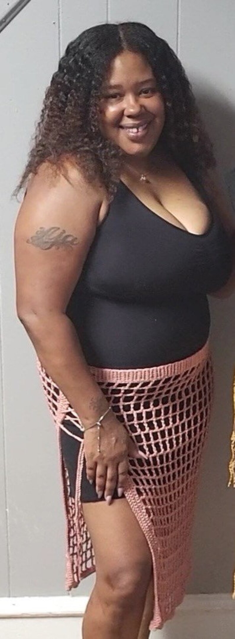 Puede incluir: Una mujer con una camiseta sin mangas negra y una falda de crochet rosa. La falda tiene un dise&ntilde;o de red. La mujer tiene el pelo rizado oscuro y un tatuaje en el brazo. Est&aacute; sonriendo.