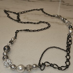 Puede incluir: Un collar plateado y negro con una cadena y cuentas. Las cuentas son una mezcla de perlas, cristales y vidrio opaco. La cadena es una mezcla de dos estilos, uno con eslabones ovalados y otro con eslabones redondos.