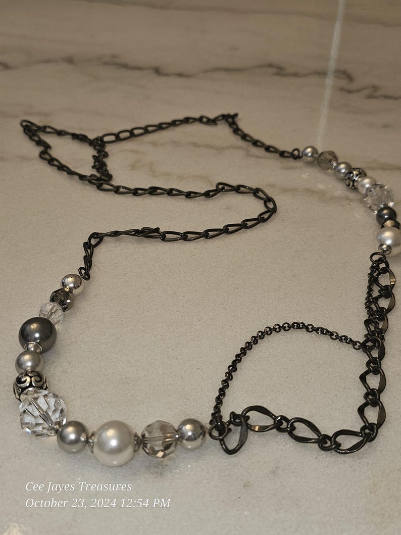 Puede incluir: Un collar plateado y negro con una cadena y cuentas. Las cuentas son una mezcla de perlas, cristales y vidrio opaco. La cadena es una mezcla de dos estilos, uno con eslabones ovalados y otro con eslabones redondos.