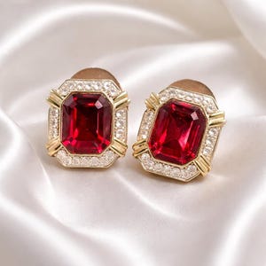 May include: A pair of gold-coloured clip-on earrings. Each earring features a large, rectangular, deep red gemstone surrounded by a border of small, clear crystals. The earrings have a gold frame with a geometric design.