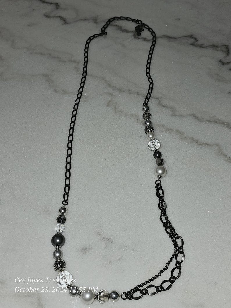 Puede incluir: Un collar de cadena negra con cuentas plateadas y blancas. El collar tiene un dise&ntilde;o de cadena doble y presenta una variedad de formas y tama&ntilde;os de cuentas.