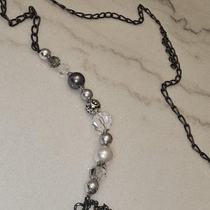 Puede incluir: Un collar de cadena plateado y negro con una hebra de cuentas plateadas y blancas. Las cuentas son una mezcla de redondas, facetadas y decorativas. La cadena tiene un cierre en forma de coraz&oacute;n.