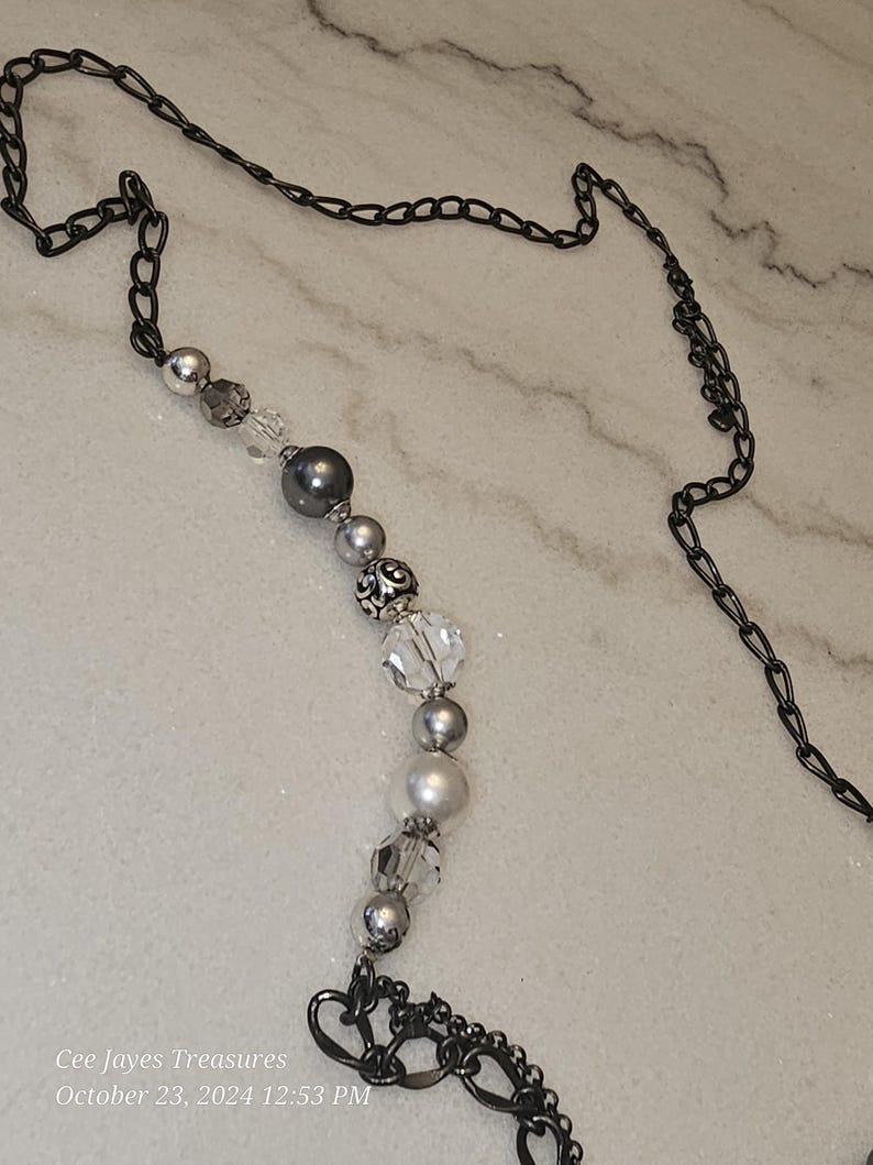 Puede incluir: Un collar de cadena plateado y negro con una hebra de cuentas plateadas y blancas. Las cuentas son una mezcla de redondas, facetadas y decorativas. La cadena tiene un cierre en forma de coraz&oacute;n.